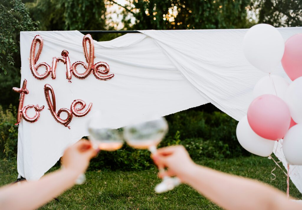 Möhippa - pynt. Två händer som skålar framför ett vitt lakan, framför är ballonger som formar texten "Bride to be"